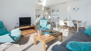 A modern living room with a TV, teal chairs, a wooden coffee table, and a dining area with white chairs and a wooden table. - Parc Océan by Interhome (Quiberon)