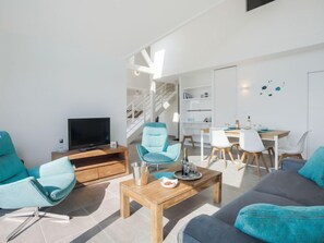 Uma sala de estar moderna com TV, cadeiras verde-água, uma mesa de centro de madeira e uma área de jantar com cadeiras brancas e mesa de madeira.