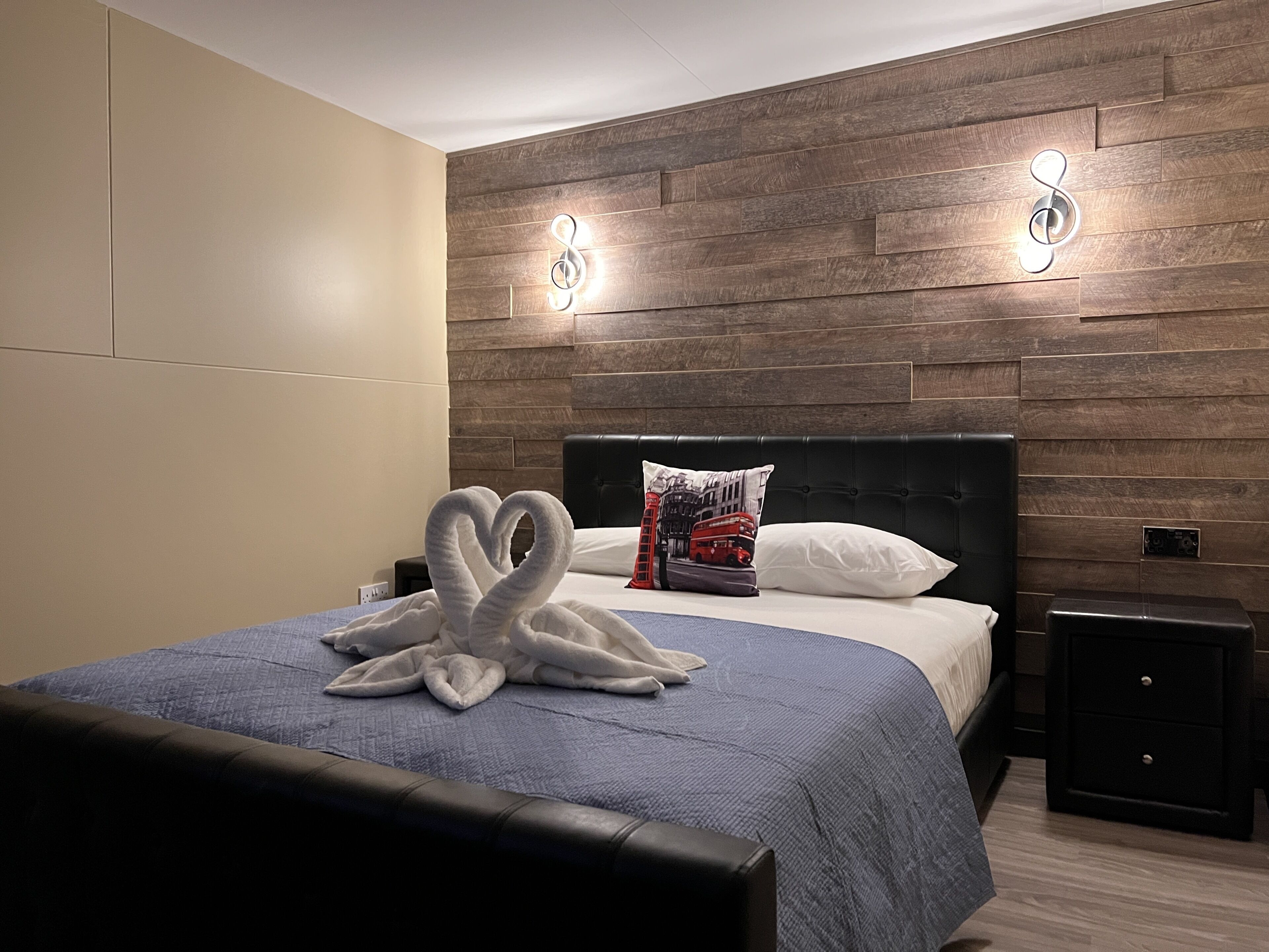 A modern hotel room with a wooden headboard, a bed with a blue blanket, a decorative towel, and a pillow with a cityscape design.