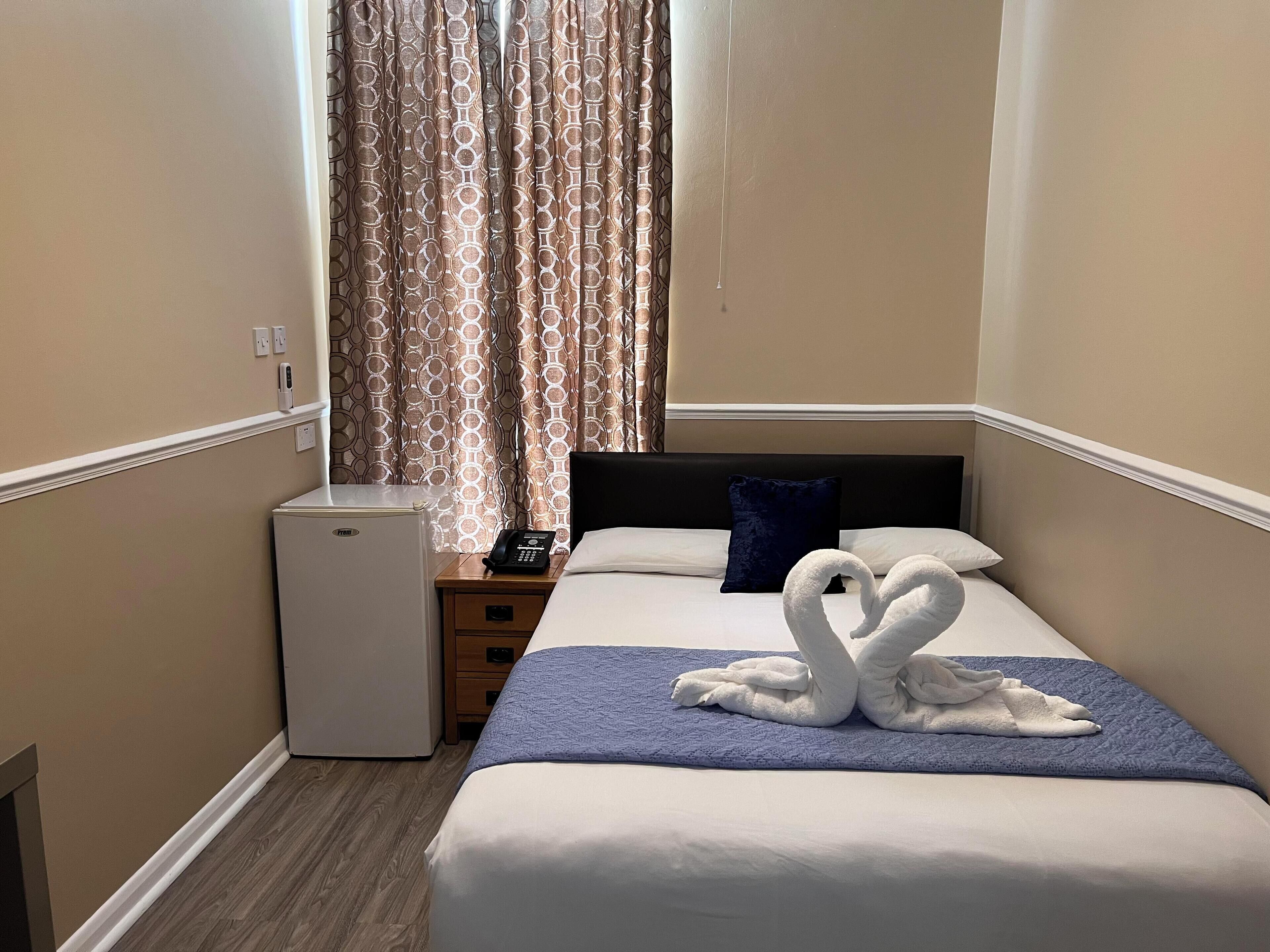 A hotel room with a bed, a small refrigerator, a bedside table with a phone, and a window with patterned curtains.