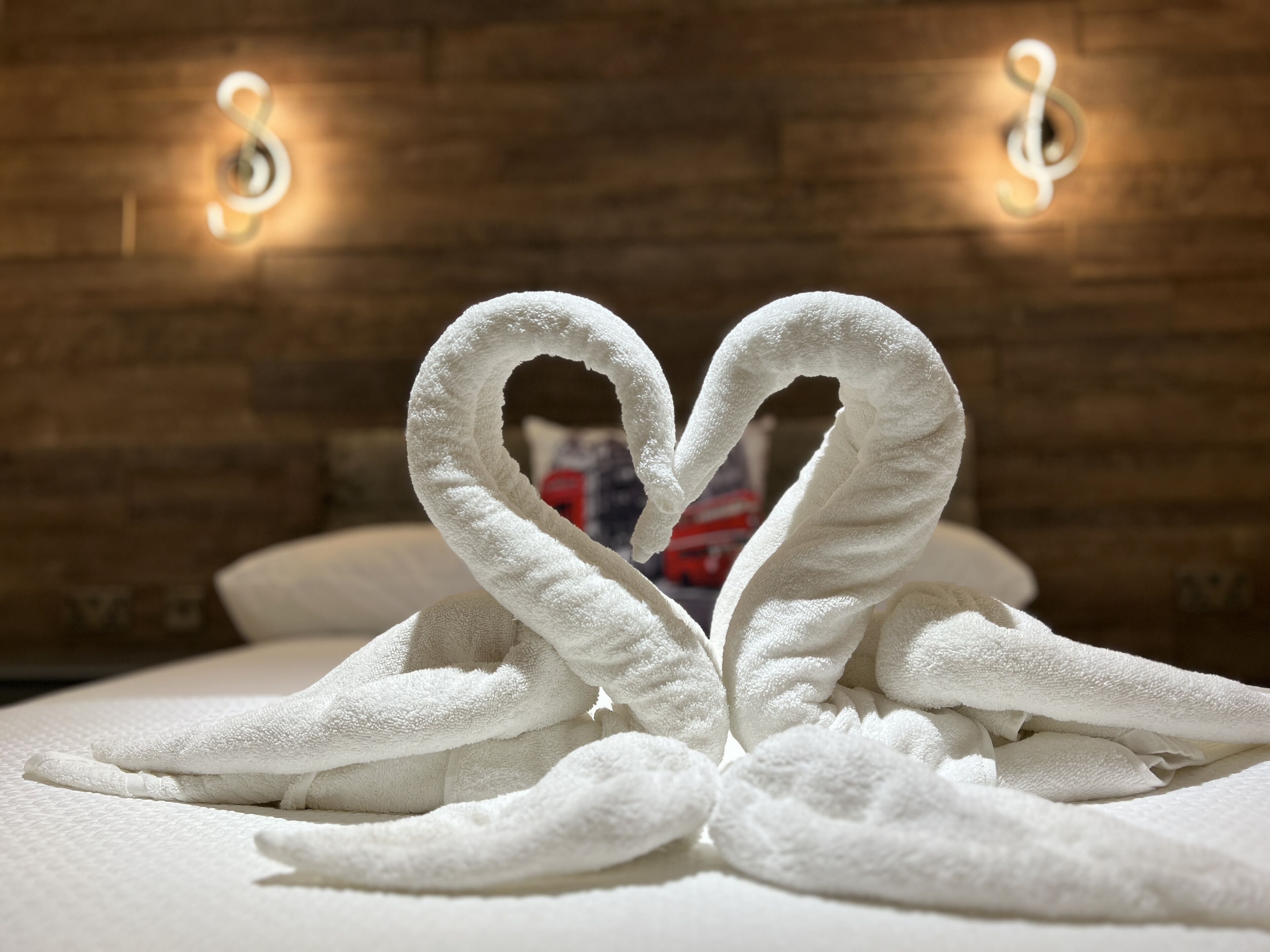 A neatly folded towel arrangement in the shape of a heart on a bed with a wooden headboard and wall-mounted lights.