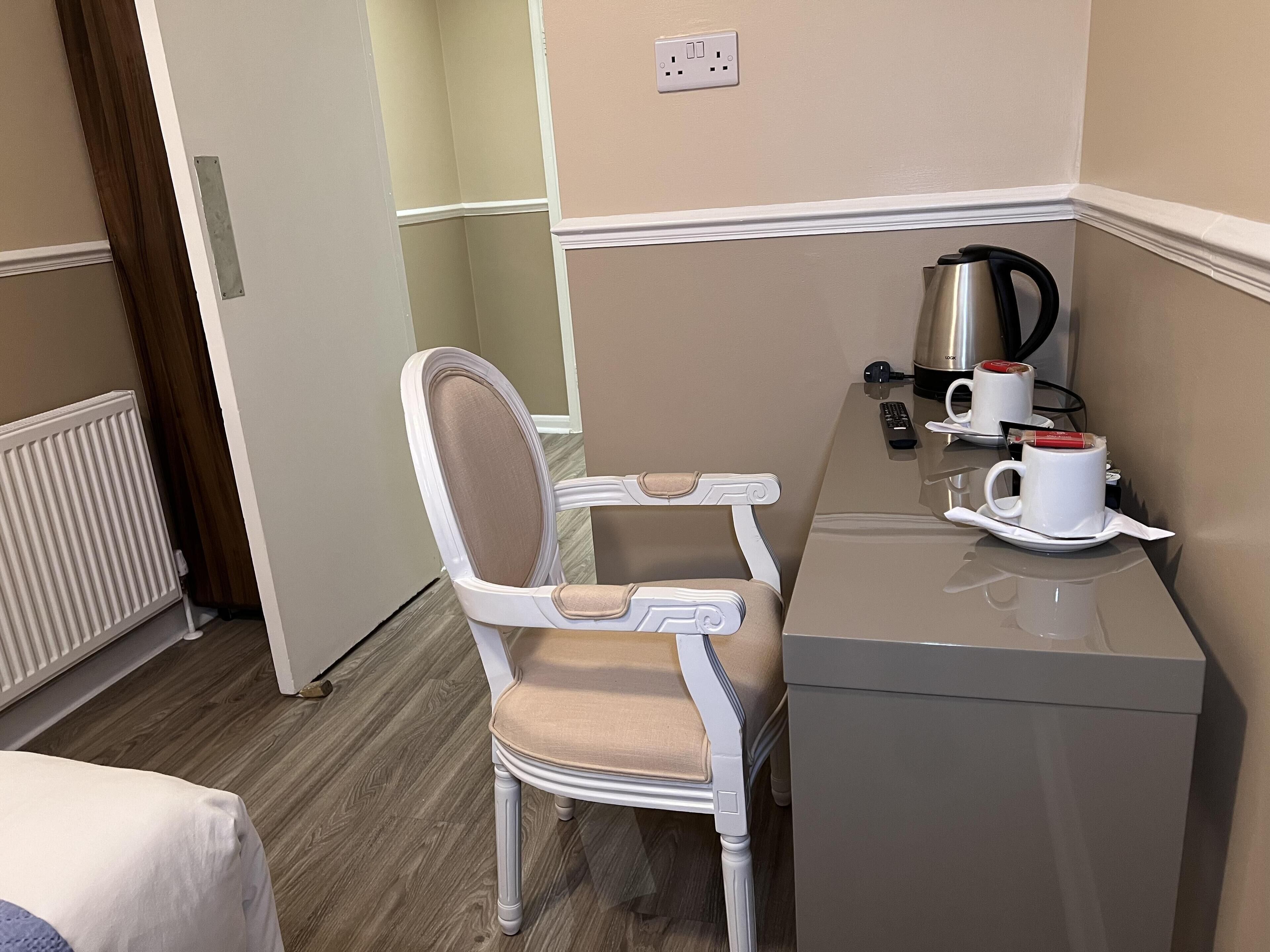 A hotel room with a white upholstered chair, a small table with a kettle and two cups, and a radiator on the floor.