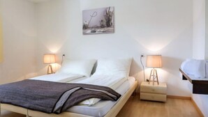 Un lit double avec une couverture grise, deux tables de chevet avec des lampes, un petit tiroir de chevet et un tableau accroché au mur.