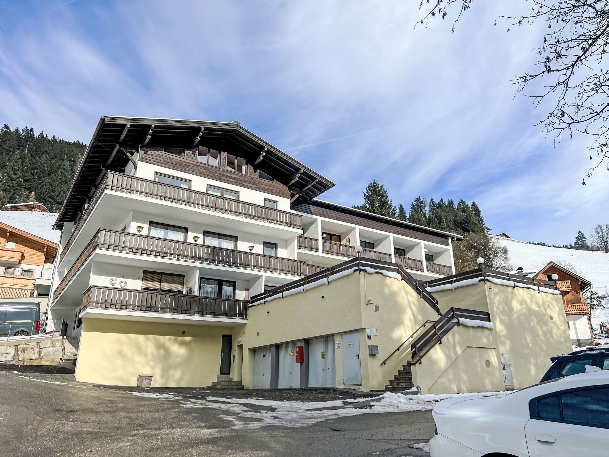 Et hotel med flere etager, altaner, parkeringsplads og et sneklædt landskab i baggrunden.
