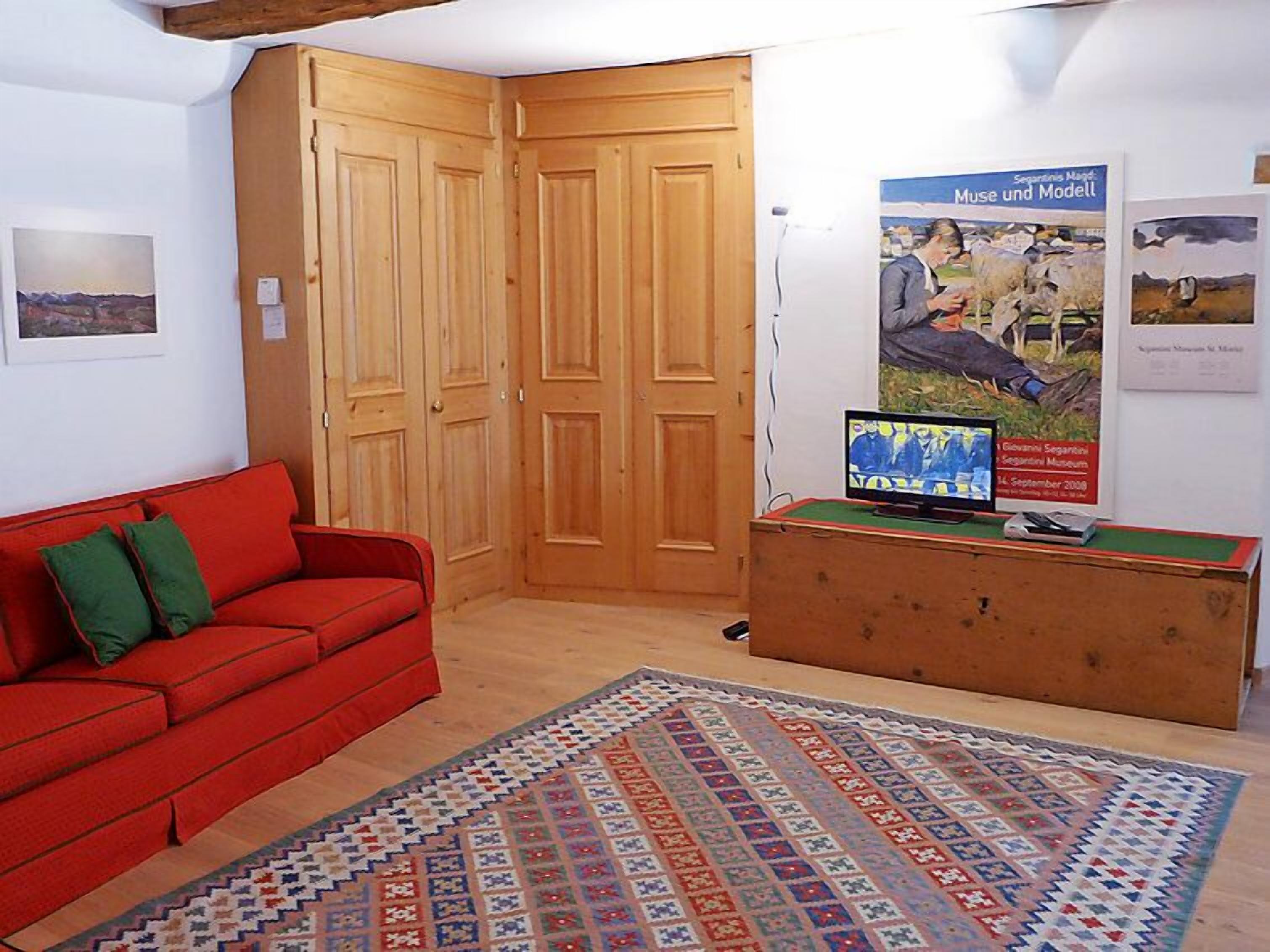 Um quarto com um sofá vermelho, um tapete estampado, portas de madeira e um pôster na parede.