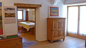 Ein Zimmer mit einem Holzkasten, einem Esstisch mit Stühlen, einem Gemälde an der Wand und Blick durch ein Fenster.