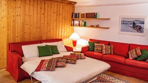Ein gemütliches Zimmer mit Holzboden, einem roten Sofa, einem Bett mit gemusterten Bettbezügen, einer Lampe, Bücherregalen und einem gerahmten Bild an der Wand.