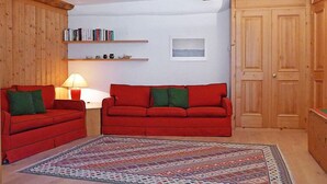 Ein gemütliches Wohnzimmer mit zwei roten Sofas, einem gemusterten Teppich, Holzregalen mit Büchern, einer Lampe und einer Holztür.