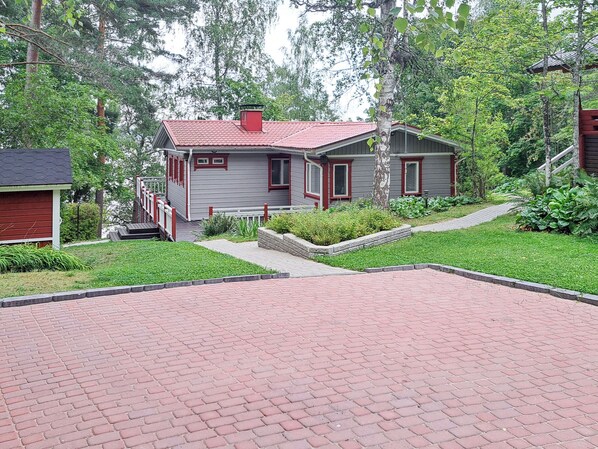 Une petite maison à un étage, avec un toit rouge et un revêtement extérieur gris, entourée d’une végétation luxuriante.