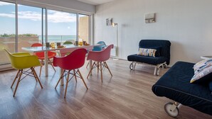 Una sala moderna con un ventanal grande con vistas al mar, un comedor con sillas coloridas y una zona de estar acogedora con un sofá azul.