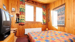 Ein gemütliches Zimmer mit Holzwänden, einem Bett mit farbenfroher Steudecke, einem Fernseher, einem Schminktisch und einem Fenster mit Vorhängen.