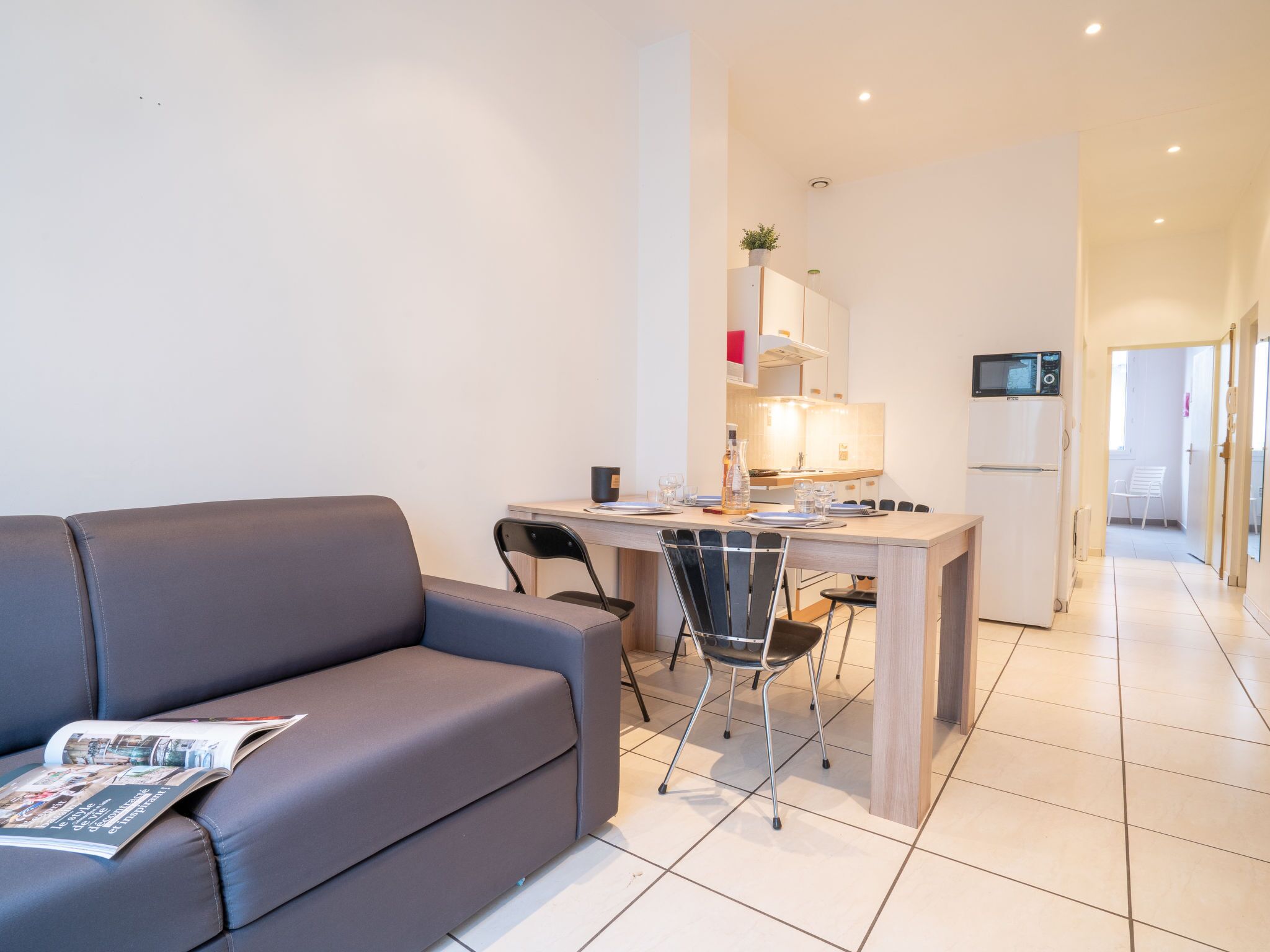A modern living room with a grey sofa, a dining table with chairs, a kitchen area with a microwave, and a refrigerator.