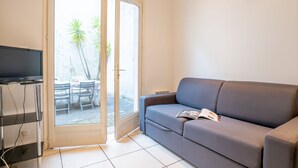 Een woonkamer met een grijze bank, een flatscreen-tv op een standaard en een schuifpui naar een buitenruimte met een tafel en stoelen.
