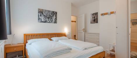 A bedroom with a wooden bed, white bedding, a nightstand, a desk, and a wardrobe.