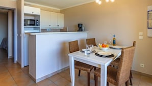 A compact kitchen with white cabinets, a microwave, and a coffee maker. A dining table with four wicker chairs is set with plates, glasses, and a fruit bowl. - Les Restanques Golfe Saint Tropez by Interhome (Grimaud)
