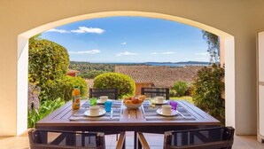 A dining table set with plates, cups, and a bowl of fruit, overlooking a scenic landscape with houses and trees. - Les Restanques Golfe Saint Tropez by Interhome (Grimaud)
