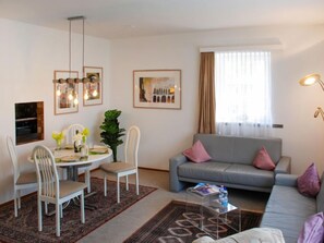 Un salon moderne avec un canapé gris, des chaises blanches, une table à manger agrémentée d’un vase de fleurs, des tableaux encadrés au mur et une grande fenêtre avec des rideaux.