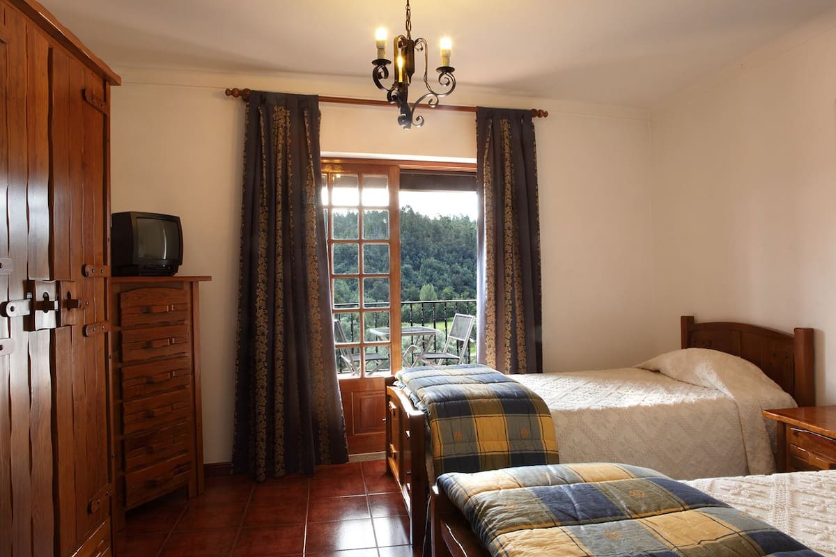 A bedroom with two beds, a wooden wardrobe, a television on a shelf, and a window with curtains.