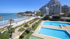 Un hotel costero con una amplia zona de piscina, acceso directo a la playa y una imponente montaña al fondo.