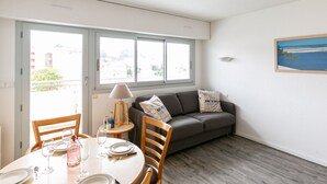 A dining area with a table set for two, a sofa, and a window with a view of buildings and trees. - Jetée des Marins by Interhome (Arcachon)
