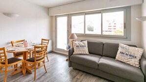 A modern living room with a grey sofa, wooden dining table, and chairs. There are two windows providing natural light and a view of buildings outside. - Jetée des Marins by Interhome (Arcachon)
