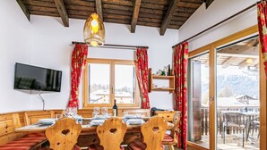 A dining area with wooden furniture, a television mounted on the wall, and a view of the outdoors through large windows. - Residenza Chesa Margun 15-5 by Interhome (Silvaplana-Surlej)