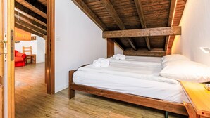 A bedroom with a wooden ceiling, a bed with white linens, a wooden nightstand, and a red chair in the adjacent room. - Residenza Chesa Margun 15-5 by Interhome (Silvaplana-Surlej)