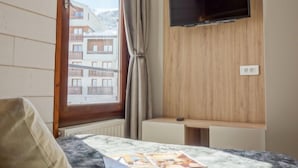 A hotel room with a bed, a flat-screen TV mounted on the wall, a window with curtains, and a view of a building outside. - Le Prariond (Val Claret) by Interhome (Tignes)