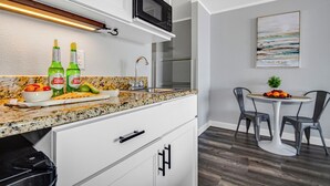 A kitchen with white cabinets, a granite countertop, a microwave, and a sink. There are two bottles of Stella Artois beer and a bowl of fruit on the counter. - Ideal Oceanview Studio Getaway! Amenities Galore (Myrtle Beach)