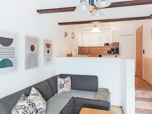Een moderne woonkamer met een grijze bank, gekleurde kussens en een houten salontafel. Achtergrond toont een keuken met een raam en een hanglamp.