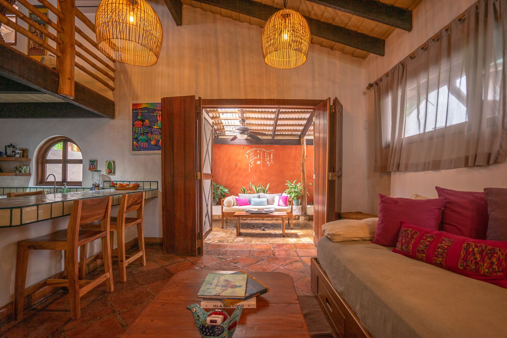 A cozy interior with a wooden dining area, a sofa with colorful pillows, and a view through an open doorway to a bright room with a ceiling fan.