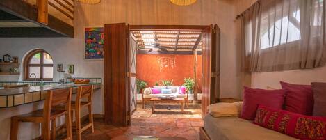 A cozy interior with a wooden dining area, a sofa with colorful pillows, and a view through an open doorway to a bright room with a ceiling fan.