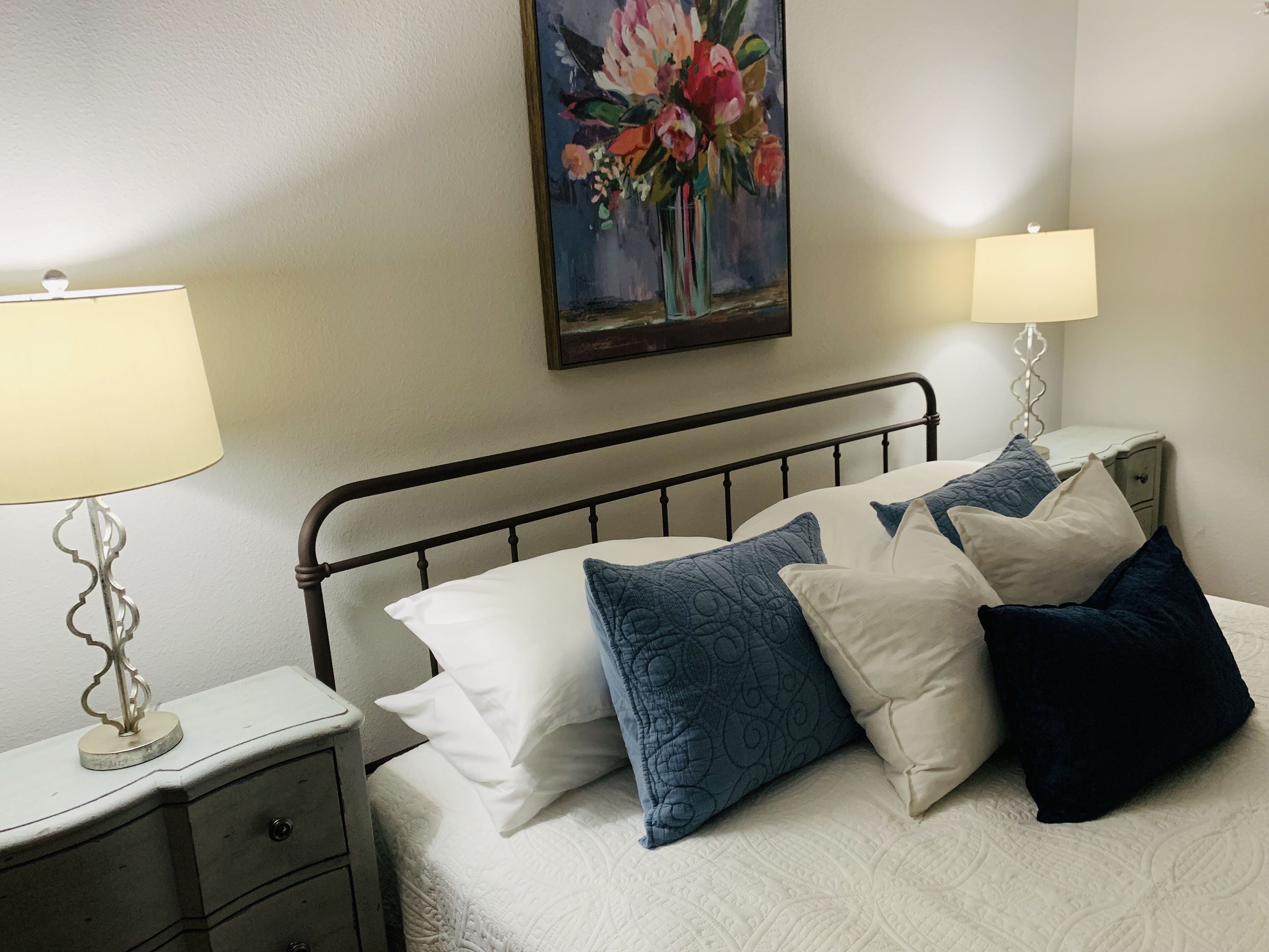 A neatly made bed with a metal frame, white bedding, and multiple pillows in shades of blue and white. There is a nightstand with a lamp and a drawer, and a painting of flowers on the wall.