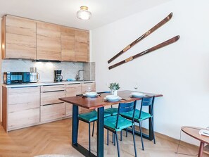 Una cocina compacta con electrodomésticos modernos, un comedor con mesa de madera y sillas azules, y esquíes colgados en la pared.