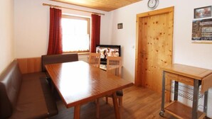 Ein gemütliches Zimmer mit Holzbalkendecke, einem Esstisch mit Stühlen, einer Couch, einem kleinen Fernseher und einer Tür mit einem Poster.