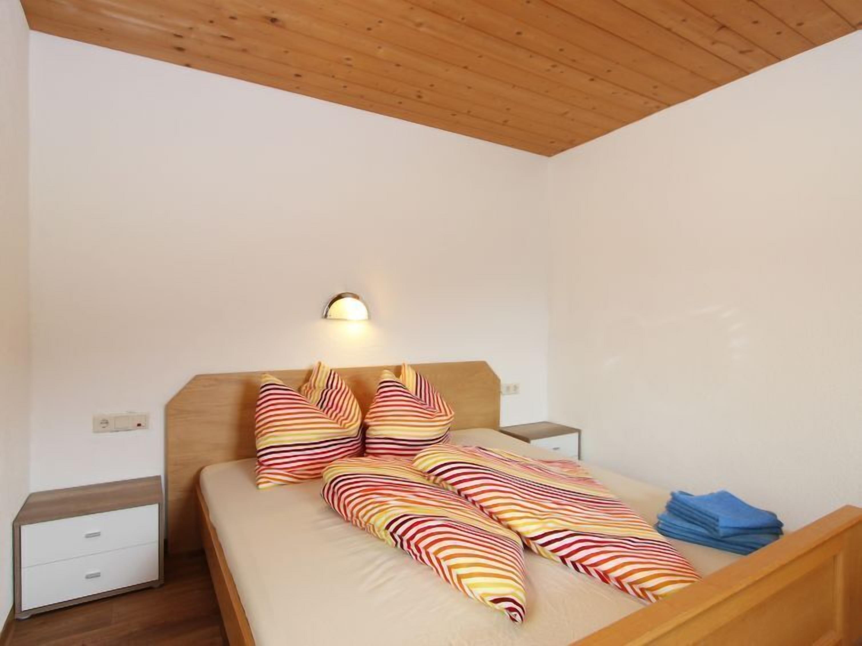 A bedroom with a wooden ceiling, a bed with striped bedding, a nightstand, and a wall-mounted light.