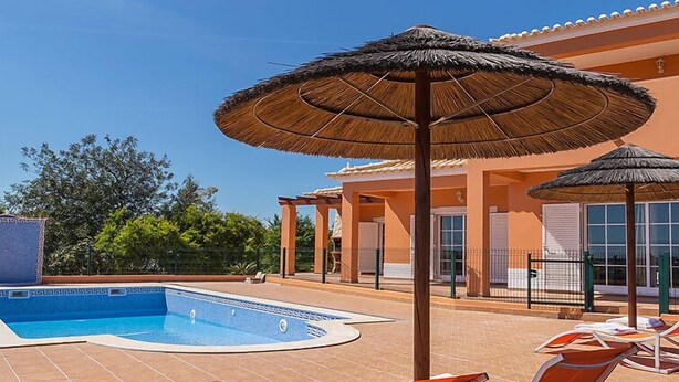A swimming pool with lounge chairs and umbrellas, surrounded by a building with a tiled roof and a fence.