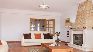 A living room with a white sofa, a wooden coffee table, a patterned rug, a stone fireplace, and a hanging light fixture.