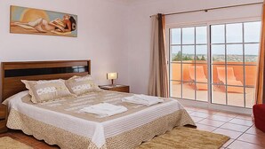 A bedroom with a large bed, a nightstand, a painting, and a view through a sliding glass door.