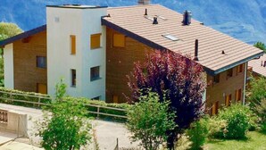 A modern two-story building with a flat roof and multiple chimneys, surrounded by lush greenery and a mountainous backdrop. - Viognier 5 by Interhome (Ovronnaz)
