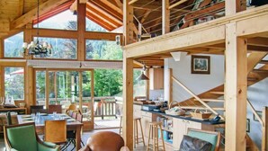 Una cabaña espaciosa de madera con comedor, dos sillones tapizados de verde, un sillón de cuero marrón y una zona de cocina con mostrador y taburetes.