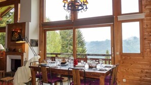 Un comedor con una mesa de madera dispuesta para cuatro personas, ventanas amplias con vistas a árboles y montañas, y una araña colgando en el techo.