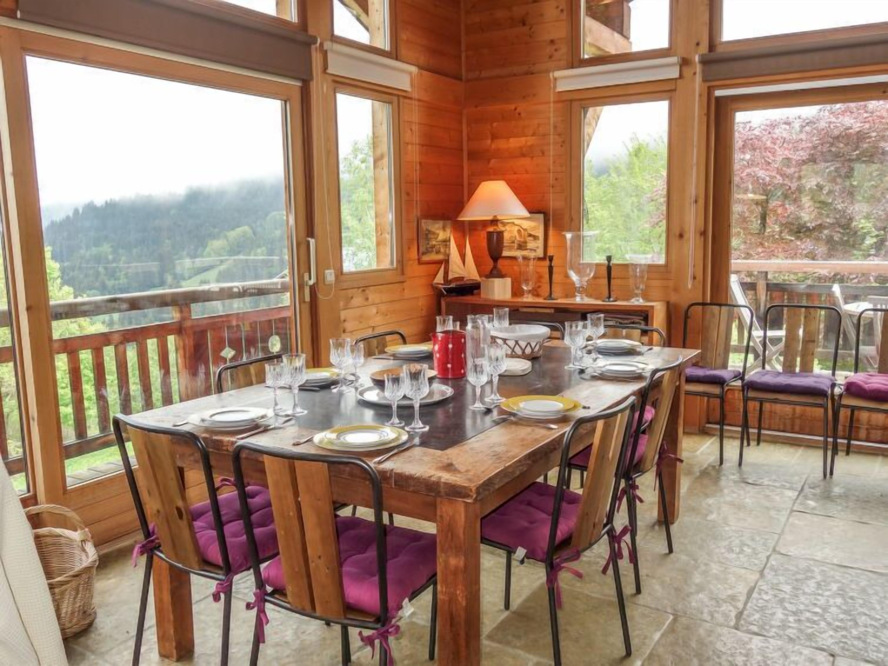 Une table à manger en bois, pouvant accueillir six personnes, entourée de chaises rembourrées de couleur violette, avec un banc en bois, et offrant une vue sur un paysage boisé grâce à de grandes fenêtres.