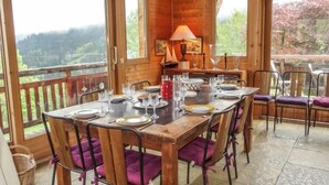 Una mesa de comedor de madera dispuesta para seis personas, con sillas con cojines morados, un banco de madera y vistas a un paisaje boscoso a través de amplias ventanas.