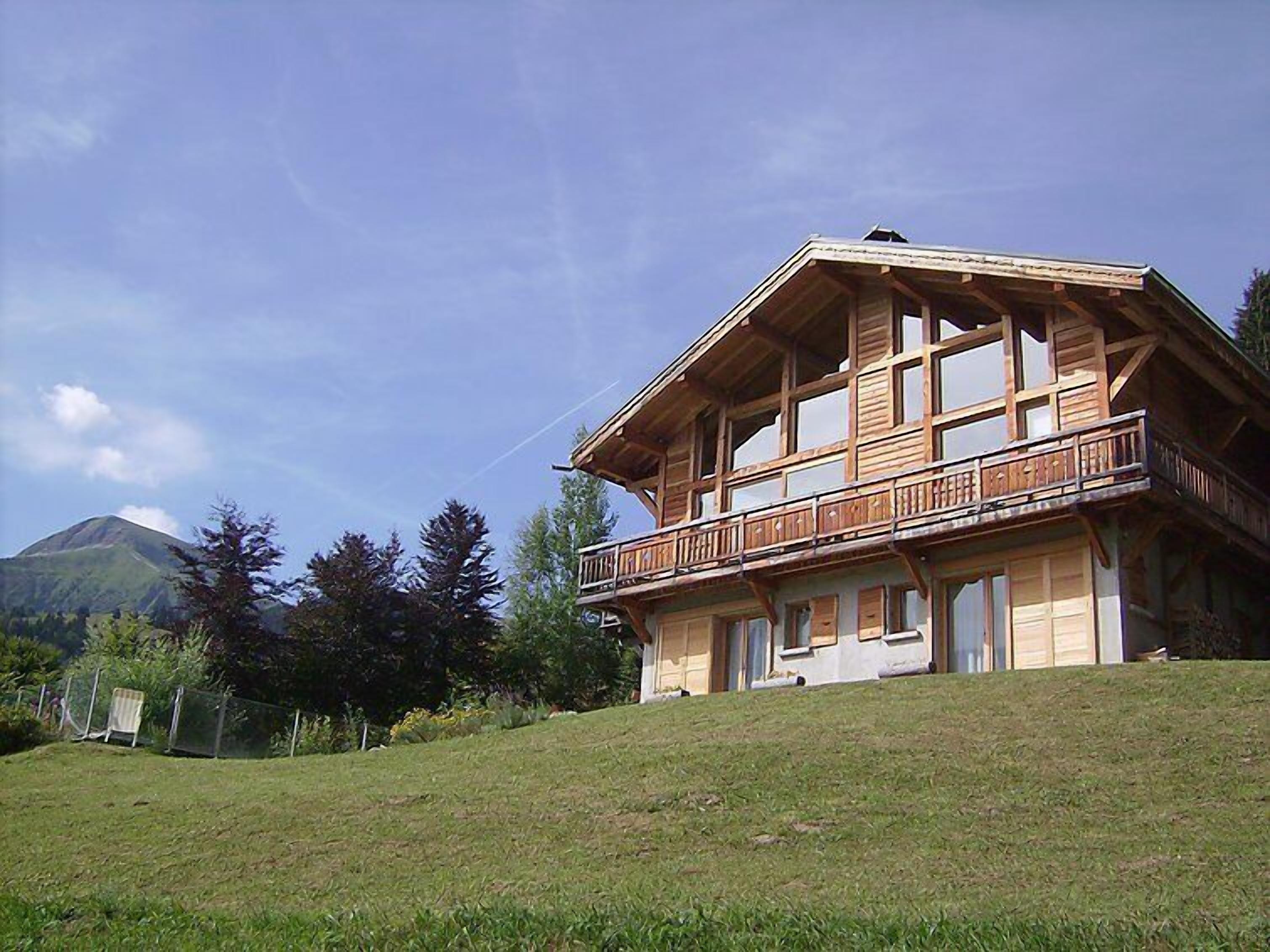 Eine zweistöckige Holzhütte mit Balkon, eingebettet in eine grüne Hanglage mit einem Berg im Hintergrund.