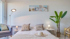 Une chambre à coucher comprenant un lit, deux tables de chevet, une lampe sur pied, une chaise, une grande plante en pot et une œuvre d’art accrochée au mur.