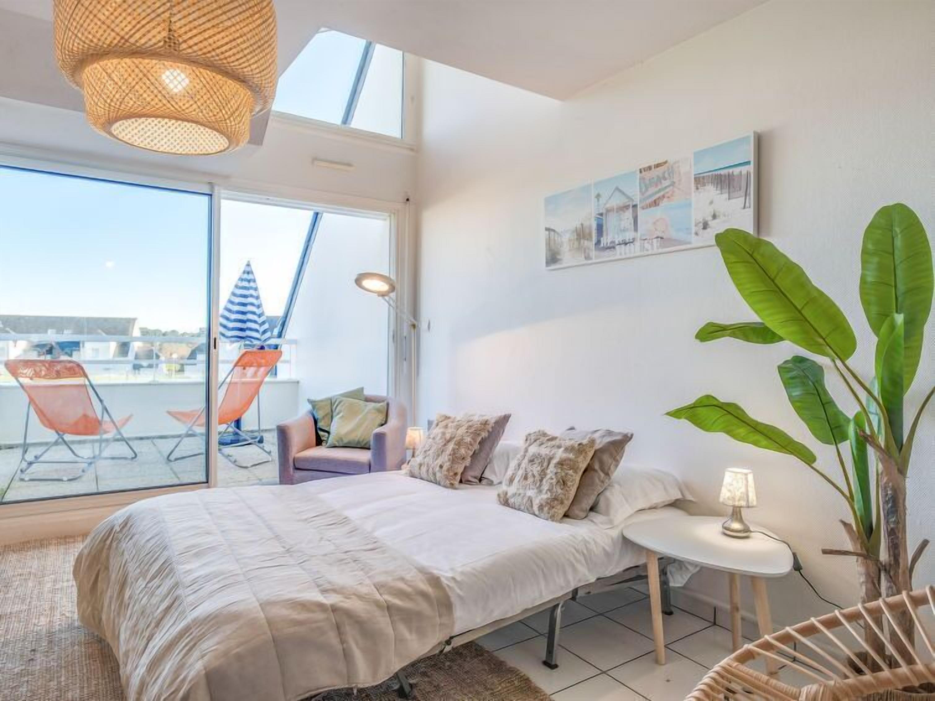 A bedroom with a bed, a small bedside table, a chair, a large potted plant, and a balcony with chairs and a striped umbrella.
