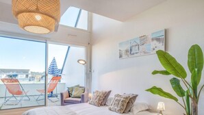 Une chambre avec un lit, une petite table de chevet, une chaise, une grande plante en pot et un balcon avec des chaises et un parasol à rayures.