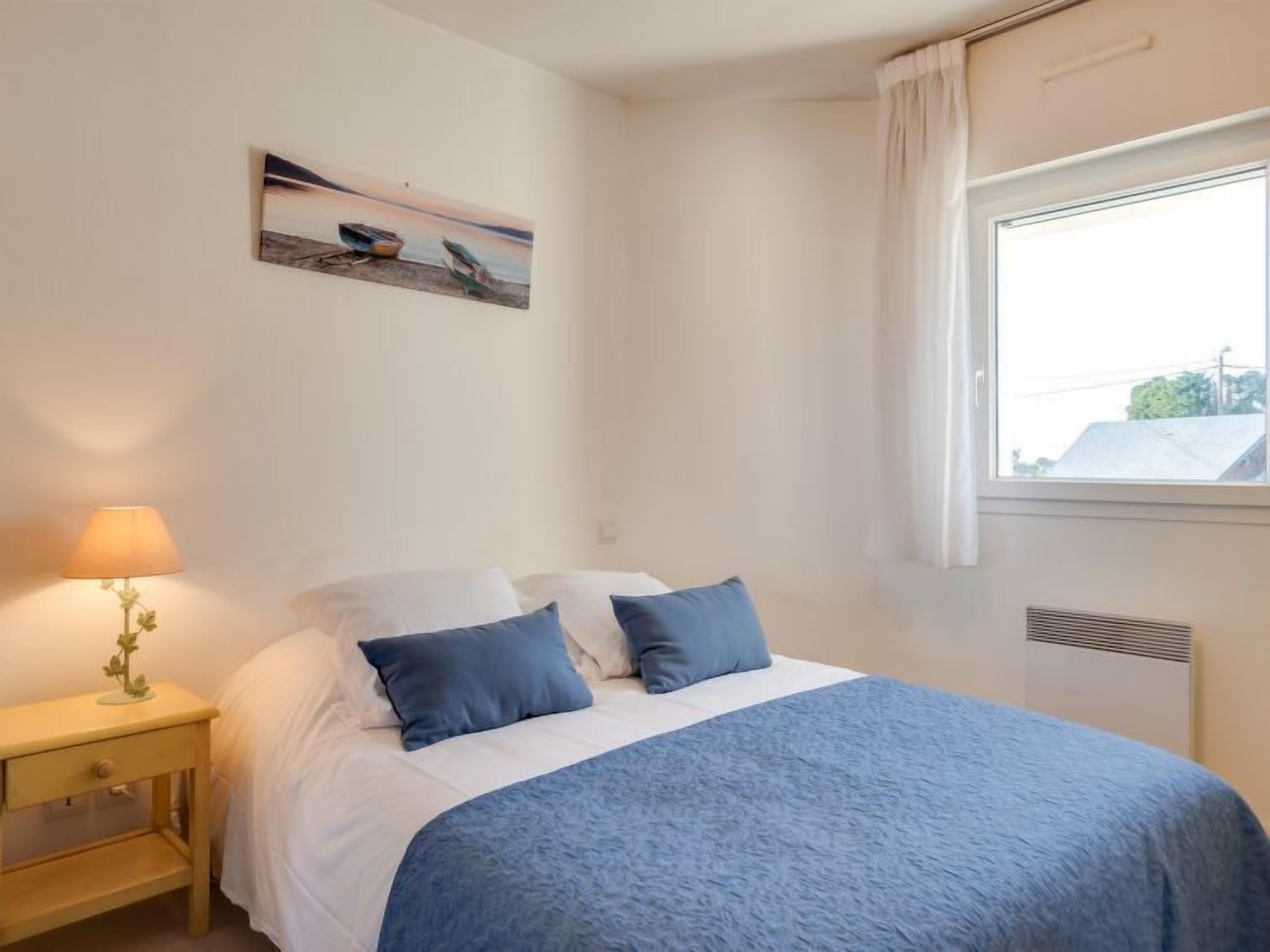 A bedroom with a bed, a nightstand, a lamp, a window, and a painting on the wall.