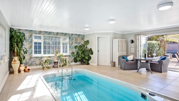 A spacious indoor swimming pool area with a clear view of the outside, surrounded by potted plants and wicker furniture. - The Coast House B&B (Bognor Regis)